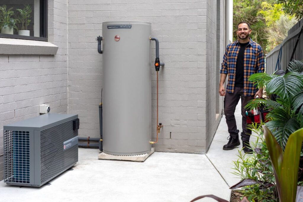 Split heat pump water heater installed outside a house: the heat pump unit on the left and the storage tank on the right, with a technician alongside
