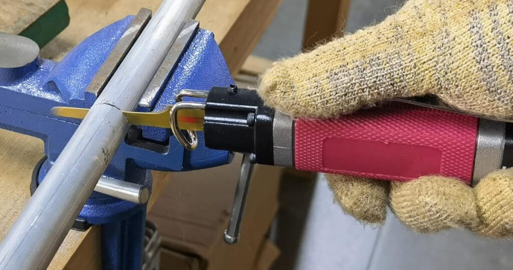 A gloved hand running a red pneumatic die grinder against a steel tube clamped in a blue bench vice, the cutting disc biting into the tube wall