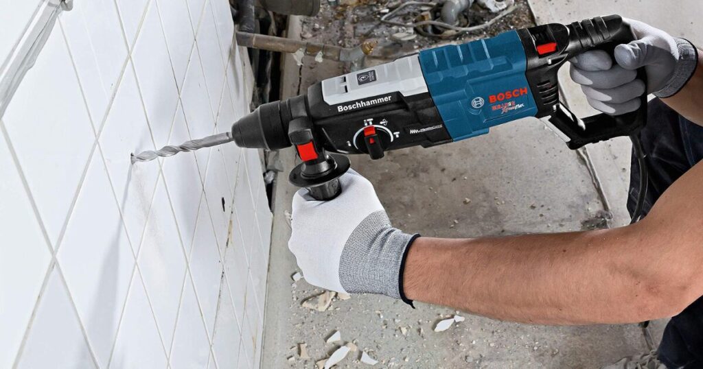 A Bosch Bulldog Xtreme SDS-plus rotary hammer drilling into a tiled wall, held with both hands on the body and the side handle, with the mode selector visible on top