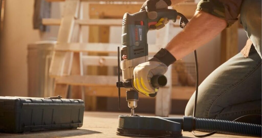 A gloved worker using a Bosch corded hammer drill with a keyed chuck, side handle and depth rod, fitted with a dust extraction shroud and hose, drilling downward into a board