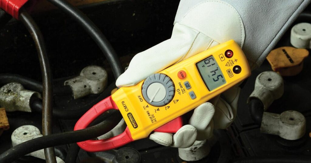 A gloved hand holding an Amprobe CM79 true-RMS clamp meter with its red jaw closed around a black cable, taking a current reading