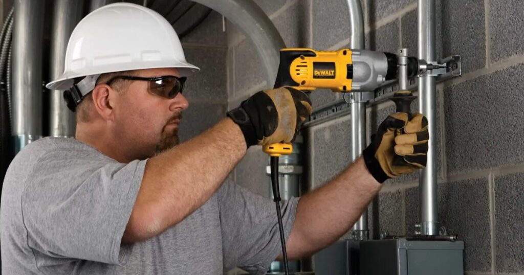 A worker in a hard hat and safety glasses using a corded DeWalt drill to fix steel conduit brackets to a concrete block wall