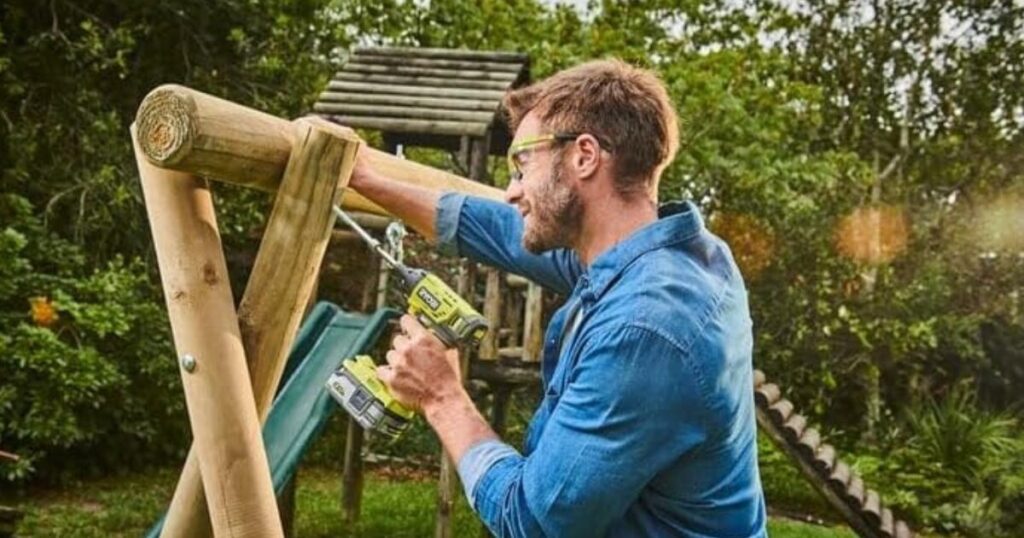 A man in safety glasses driving a screw into a timber garden climbing frame with a Ryobi cordless drill driver