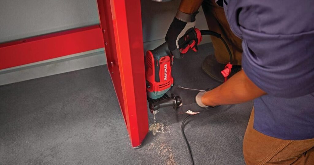A gloved worker using a red Craftsman corded hammer drill with side handle to drill a fixing into a concrete floor beside a cabinet