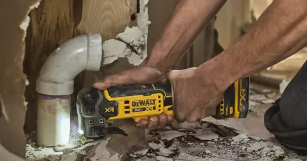 A DeWalt 20V MAX XR cordless oscillating multi-tool cutting into plasterboard beside a white PVC waste pipe, its work light on the cut