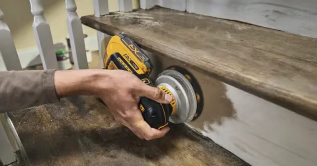 A DeWalt 20V MAX XR cordless random orbit sander being used one-handed to sand paint from a timber stair stringer