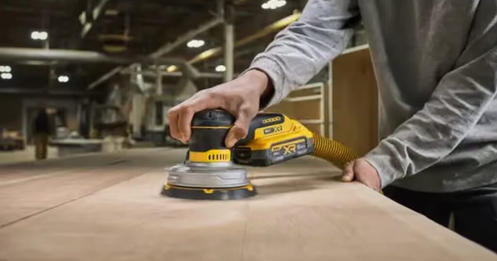 A DeWalt 20V MAX XR cordless orbital sander with a dust extraction hose attached, sanding a large plywood panel in a workshop