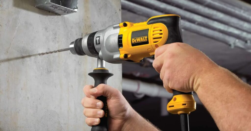 A corded DeWalt hammer drill with its side handle fitted, driving a masonry bit into a concrete wall