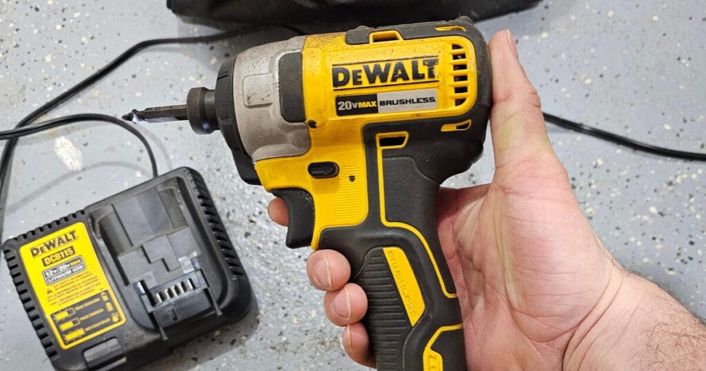 A well-used DeWalt 20V MAX brushless impact driver held in one hand with a bit seated in its quarter-inch hex collet, a DeWalt DCB115 charger on the floor beside it
