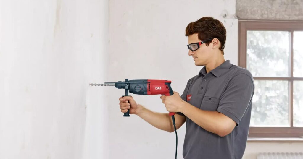 A worker in safety glasses using a red FLEX corded SDS rotary hammer with side handle to drill into a plastered wall