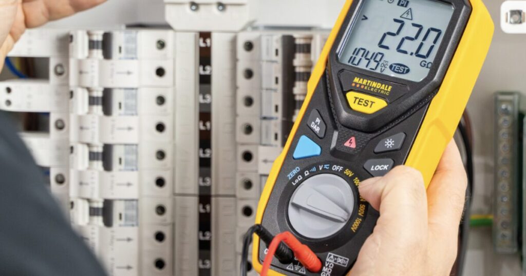 An electrician holding a Martindale insulation tester showing a 22.0 gigaohm reading in front of an open distribution board full of MCBs