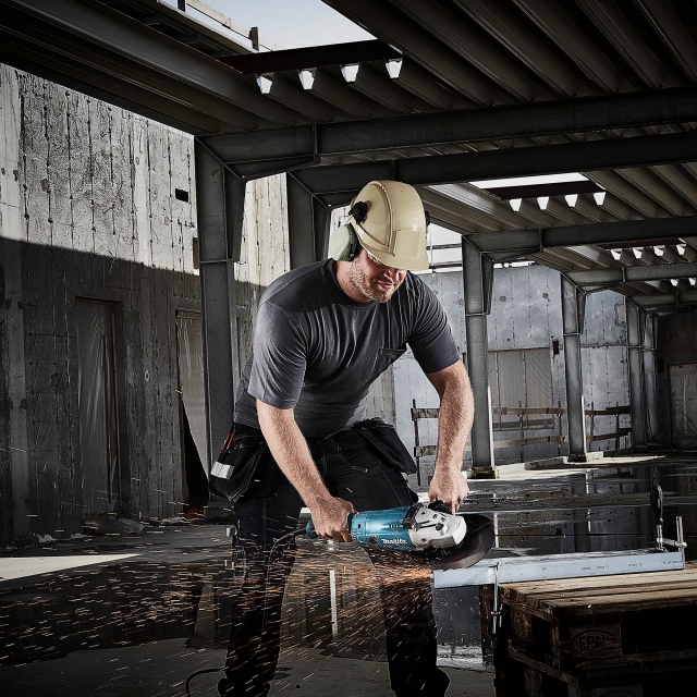 A worker in a hard hat and ear defenders cutting steel with a Makita angle grinder, sparks flying, on an unfinished steel-framed building