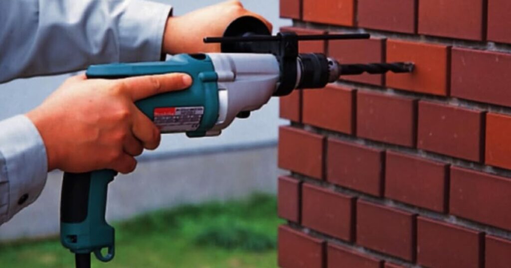 A teal Makita corded hammer drill with side handle and depth rod drilling into a red brick wall