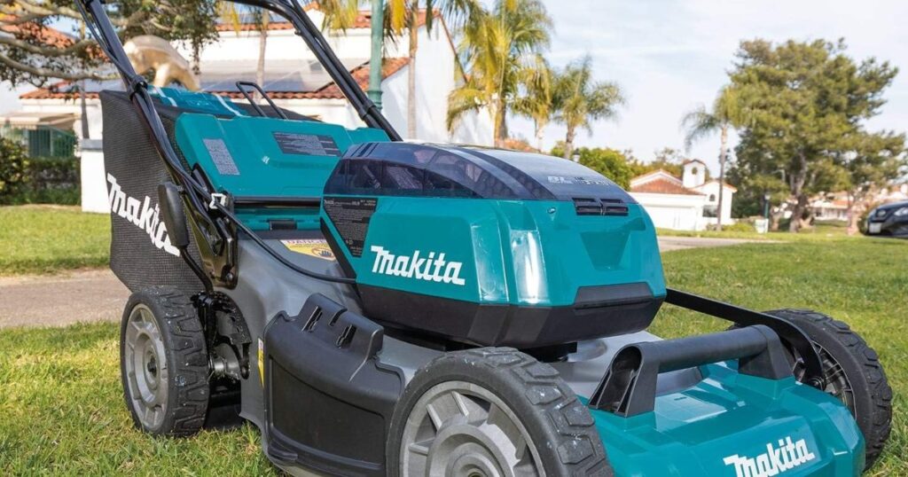 A Makita cordless lawn mower with its grass collection bag fitted, standing on a cut lawn in front of a suburban house
