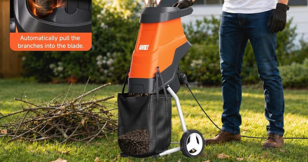 An operator in work gloves feeding branches into an orange electric chipper shredder with a mesh collection bag, a pile of cut branches alongside