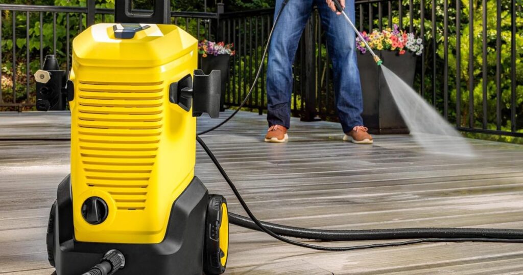A yellow electric pressure washer standing on a timber deck with its hose run out, a person spraying the deck boards behind it