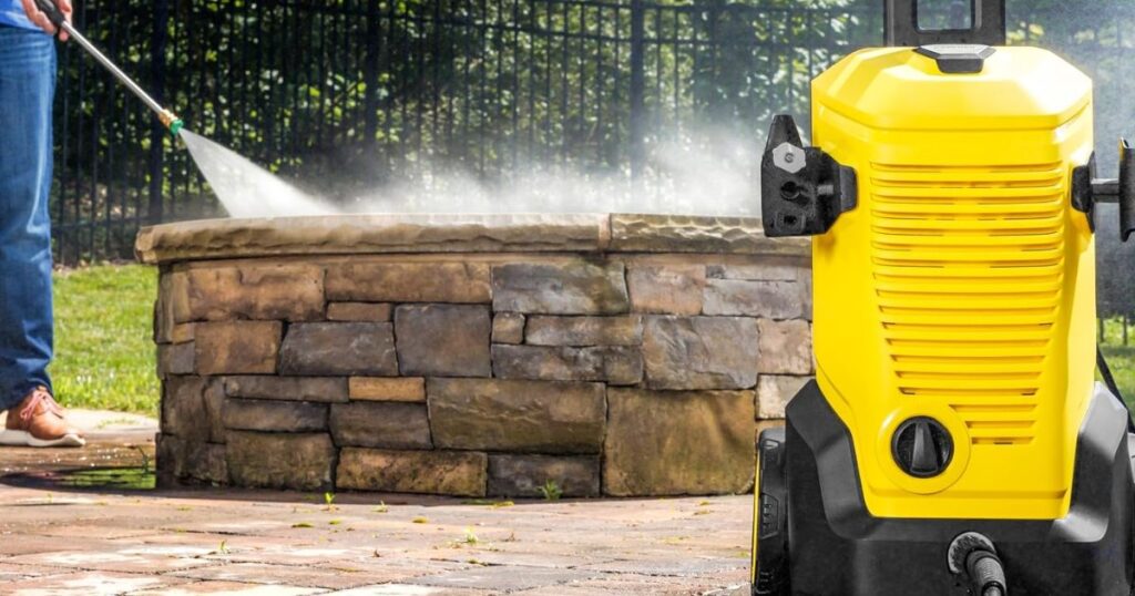 A person using an electric pressure washer to clean a stone garden wall, the spray plume visible and the yellow machine standing in the foreground