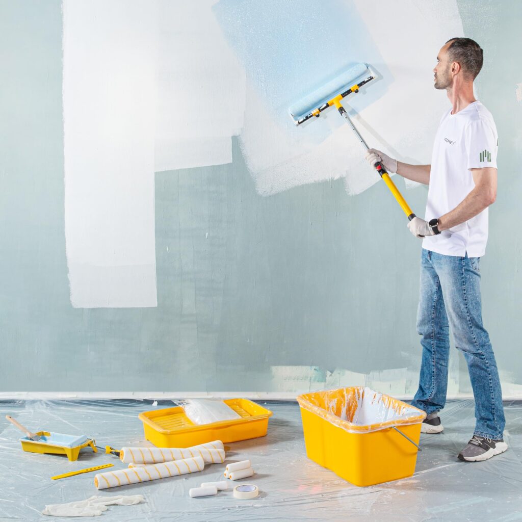 A decorator rolling a pale blue coat onto a large interior wall using a wide roller on an extension pole, with trays, mini roller covers and a paint bucket on the dust sheet
