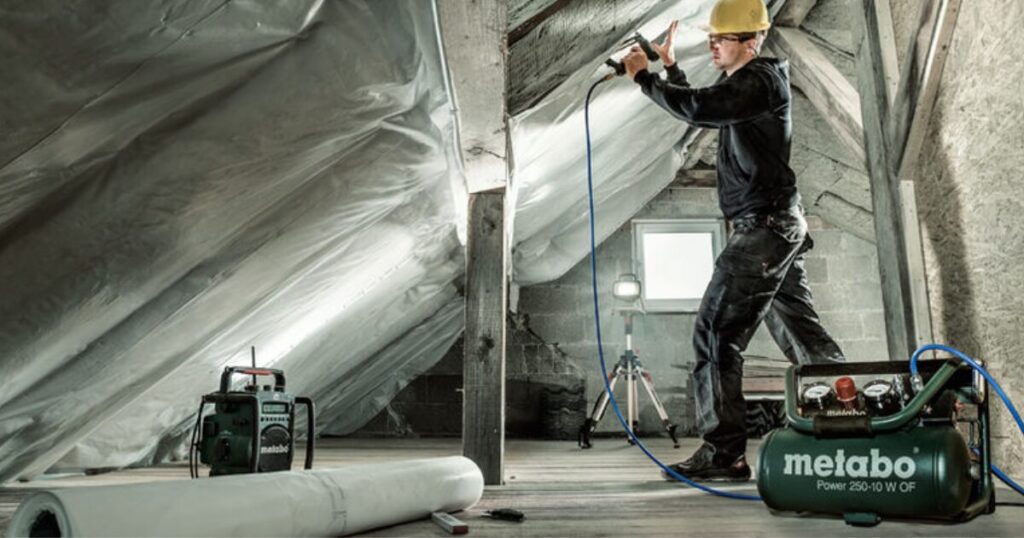 A worker in a hard hat firing a pneumatic nailer overhead into roof timbers in a loft, with a blue air hose running down to a Metabo portable compressor on the floor