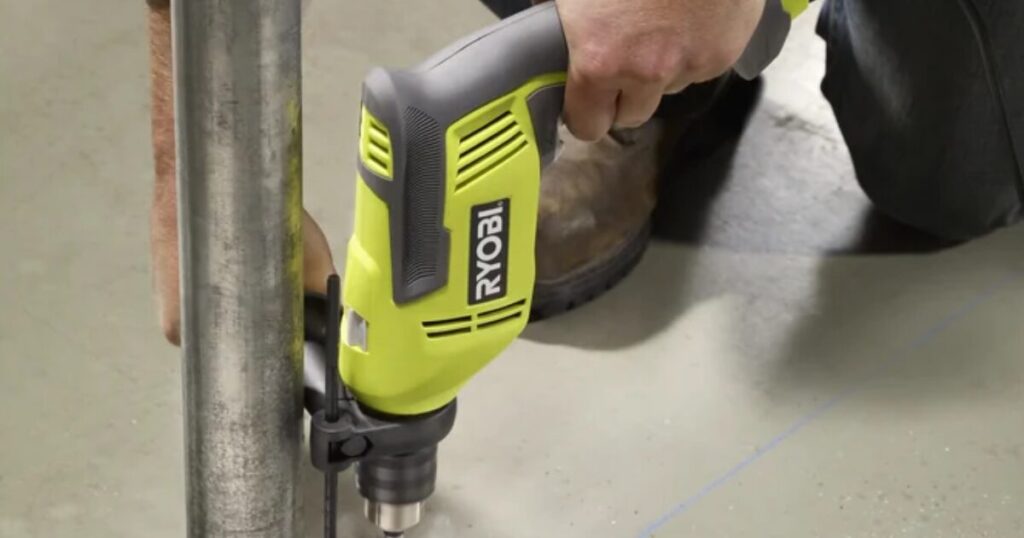 A green Ryobi corded hammer drill with a keyed chuck and chuck key fitted, drilling into a concrete floor next to a steel pipe