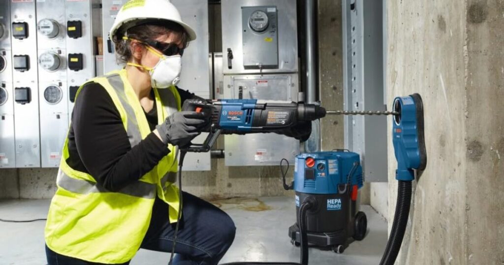 A worker in a hard hat, safety glasses, dust mask and hi-vis vest drilling a concrete wall with a corded Bosch SDS-plus rotary hammer fitted with a dust extraction shroud, a HEPA vacuum beside her