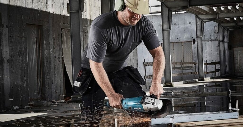 A worker in a hard hat and ear defenders cutting steel with a Makita angle grinder, sparks flying, on an unfinished steel-framed building