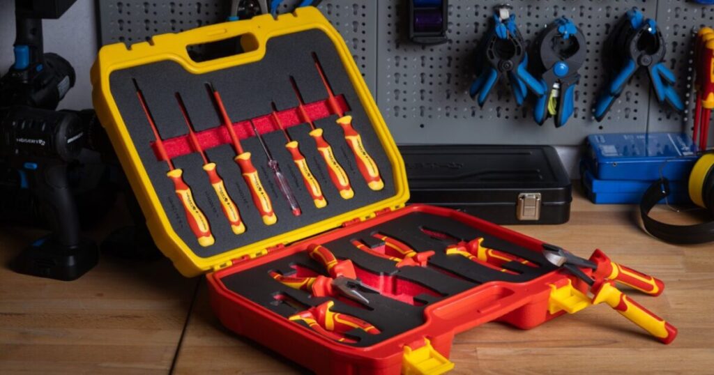 An open yellow and red case of VDE insulated screwdrivers and pliers with red and yellow handles, on a workbench in front of a tool pegboard