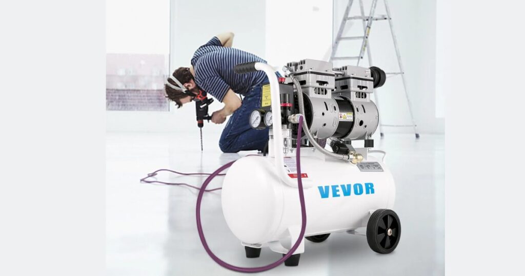 A white VEVOR oil-free twin-motor air compressor with wheeled horizontal tank and dual pressure gauges, an air hose running to a worker drilling a floor behind it