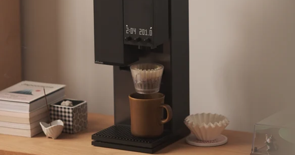 A black xBloom Studio on a wooden shelf brewing through a glass dripper into a brown mug, its LED display reading 2:04 and 201.0