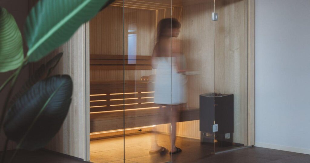 Modern indoor sauna with a glass front, two-tier timber benches lit by LED strips, and a wall-mounted heater topped with stones