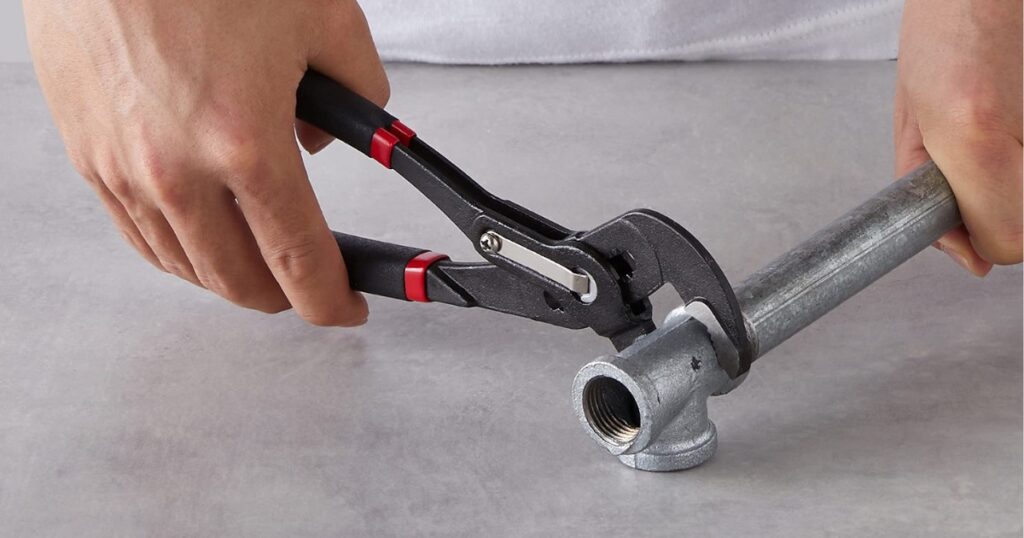 Two hands using adjustable pliers with red-trimmed handles to grip a galvanised steel pipe tee fitting
