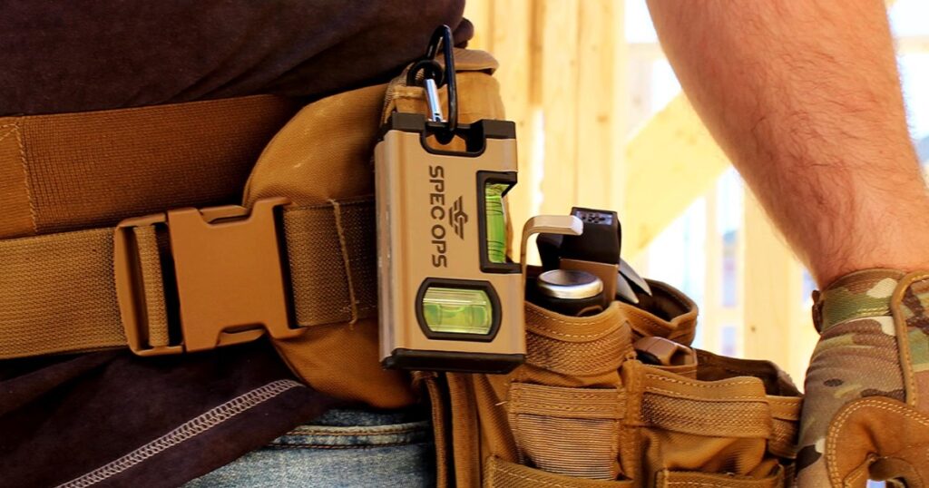 A compact torpedo level with two green vials clipped by a carabiner to a tan tool belt