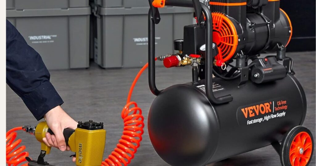 A black VEVOR oil-free air compressor with a wheeled horizontal tank and twin-cylinder pump, connected by a coiled orange hose to a pneumatic nailer held in a worker's hand
